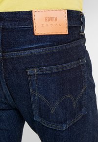Vaqueros de mezclilla azul oscuro con un parche de cuero EDWIN en la cinturilla trasera y costuras amarillas. Presenta un bolsillo trasero con costuras decorativas.