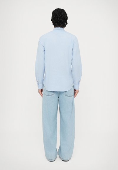 Cămașă bleu cu nasturi, mâneci lungi, tiv curbat și nasture accent roșu. Asortată cu blugi largi de culoare bleu deschis.