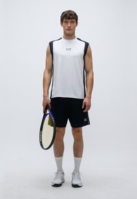 Biele atletické tielko bez rukávov s námorníckymi akcentmi, v kombinácii s čiernymi šortkami. Drží tenisovú raketu, nosí biele ponožky a tenisky.