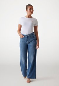T-shirt bianca aderente con un piccolo logo sul petto, abbinata a jeans blu a vita alta e gamba larga. I jeans presentano un lavaggio chiaro e tasche.
