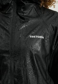 Vattentät svart jacka med en glänsande textur, med synliga vattendroppar och en vit "TRETORN"-logotyp på bröstet.