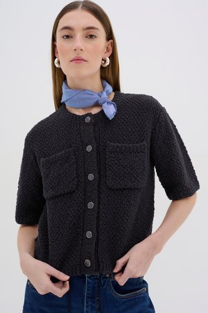Schwarze strukturierte Kurzarm-Cardigan mit rundem Ausschnitt, vorderen Knöpfen und zwei Brusttaschen. Getragen mit einem blau gestreiften Schal.