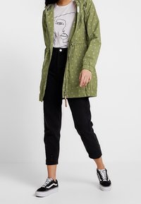 Green parka jacket with white circular pattern, adjustable hood, front pockets, paired with black cropped jeans and black sneakers.