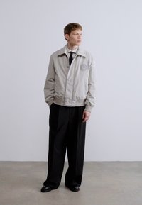 Ανοιχτό γκρι bomber μπουφάν με διακριτικό στο στήθος, ριπ μανσέτες και όρθιο γιακά, συνδυασμένο με μαύρο παντελόνι με φαρδιά πόδια.