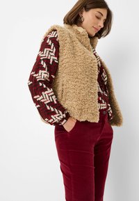 Beige Kunstfell-Weste über einem gemusterten bordeauxroten und cremefarbenen Langarmshirt, kombiniert mit bordeauxroten Cordhosen.
