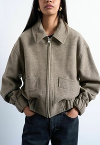 Femme portant une veste taupe à fermeture éclair avec un large col, des poignets élastiques et des poches à rabat à l'avant, associée à un jean en denim foncé. Mains dans les poches.