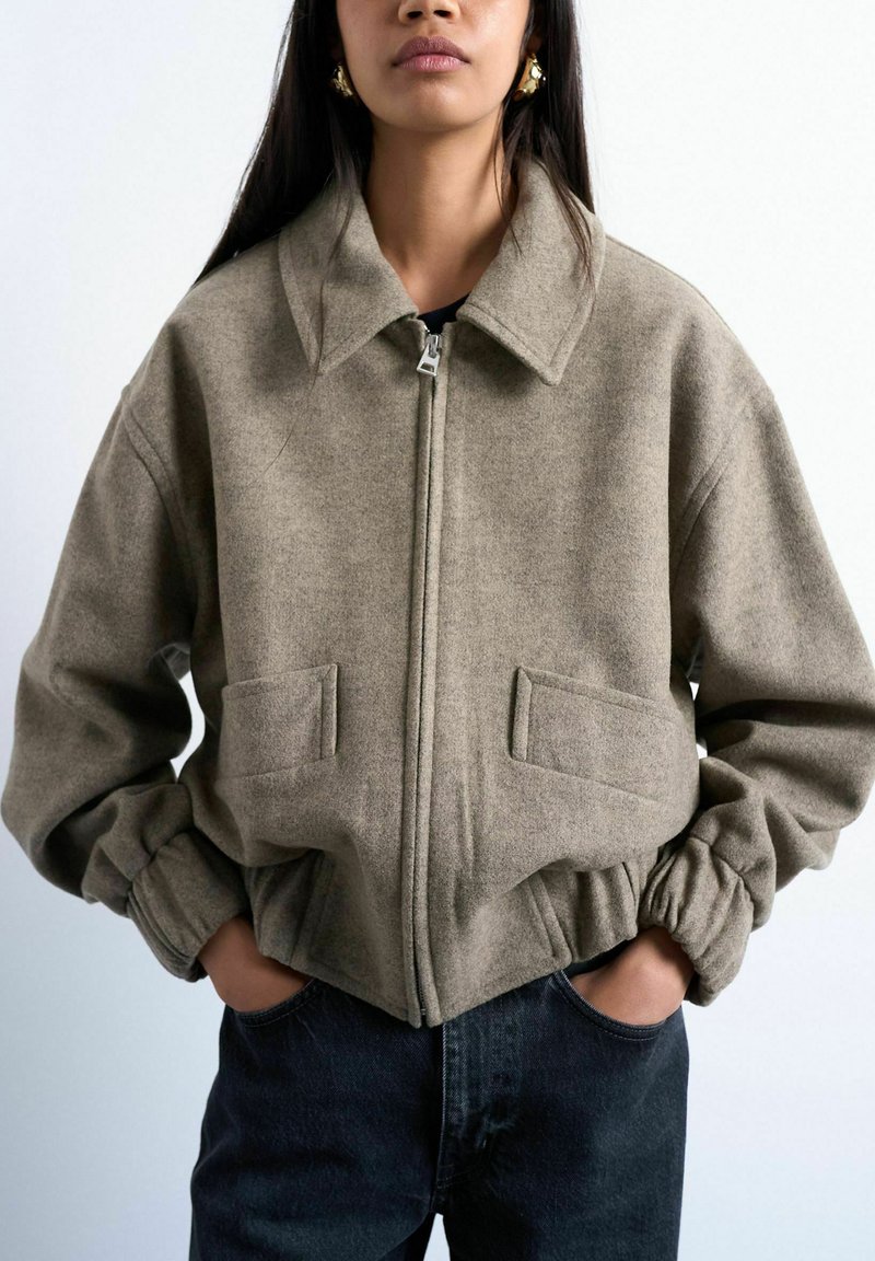 Femme portant une veste taupe à fermeture éclair avec un large col, des poignets élastiques et des poches à rabat à l'avant, associée à un jean en denim foncé. Mains dans les poches.