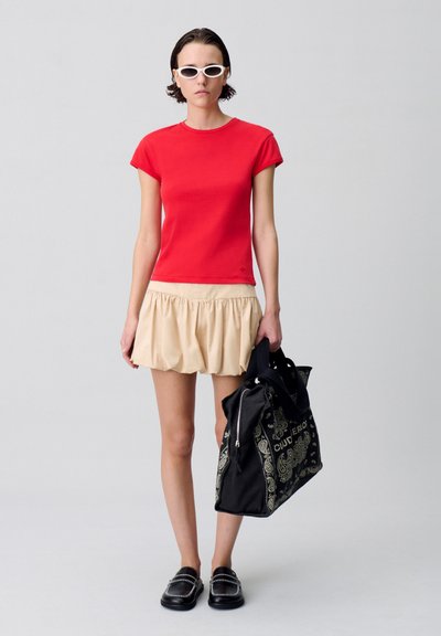 T-shirt rosso aderente a maniche corte, gonna beige con volant, scarpe nere basse con dettagli metallici e una grande borsa nera con stampa paisley bianca.