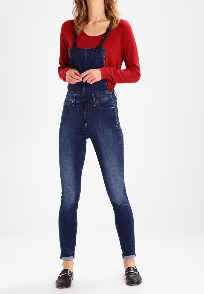 Femme portant une salopette en denim bleu foncé avec les ourlets retroussés, un chemisier rouge à manches longues et des mocassins noirs sans lacets, debout devant un fond blanc.