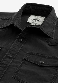 Čierna denimová košeľa s gombíkovou chlopňou a golierom, na ktorej je biely štítok s nápisom "Premium Grade 1982 Next Durable Goods."