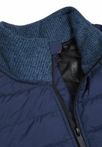 Marineblå quiltet jakke med en mørkeblå strikkrør, glat indvendig foring og en sort lynlås. Teksturen varierer mellem sektionerne.