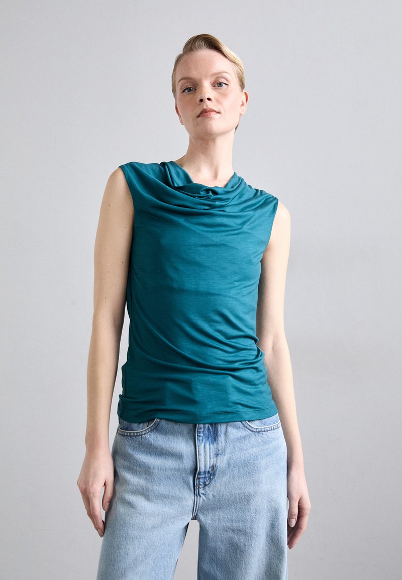 Akvamarinblå, ermeløs topp med drapert hals, laget av mykt stoff, parret med lyseblå, behagelig passform jeans. Glatt tekstur, uten mønstre.