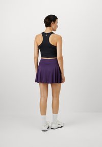 Crni crop top s racerback izrezom uparen s ljubičastom suknjom skater. Ima glatku teksturu i prilagođen siluetu. Nošeno s bijelim sportskim tenisicama.