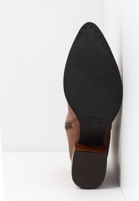 Semelle en caoutchouc noir avec une adhérence texturée et tige en cuir marron sur une botte pour femme arrivant à la cheville, posée contre une surface blanche.