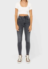 Jeans in denim nero a vita alta, dal taglio slim e design classico a cinque tasche, abbinati a una maglietta corta bianca e sneakers marroni.