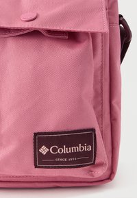 Υφασμάτινη τσαντάκι ταχυδρόμου Columbia σε ροζ αποχρώσεις με κλείσιμο με καπάκι, διαθέτοντας τσόχα με λογότυπο, υφή υλικού και κομψό σχεδιασμό.