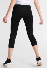 Leggings capri neri realizzati in tessuto elasticizzato, caratterizzati da una texture liscia, design aderente e lunghezza alla caviglia, abbinati a scarpe sportive nere.