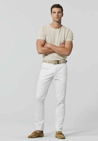 T-shirt beige à col rond, pantalon blanc à jambes droites et mocassins en suède marron. Un homme se tient avec les bras croisés, mettant en valeur une coupe décontractée et des couleurs neutres.