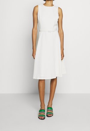 Femme portant une robe midi blanche sans manches ceinturée avec des sandales à bouts ouverts rayées vert, noir et beige, debout sur un fond blanc uni.