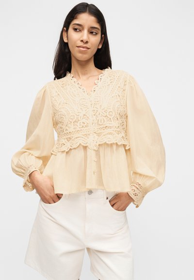 Νεαρή γυναίκα που φοράει μια μπλούζα κρεμ με λεπτομέρειες από δαντέλα και φουσκωτά μανίκια, μπουκαρισμένη σε λευκό σορτς ψηλής μέσης, τα χέρια στις τσέπες, ουδέτερο φόντο.