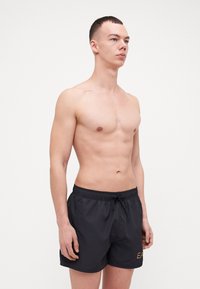 Șorturi de înot negre cu talie elastică, șnur de strângere și detaliu cu text auriu pe lateral. Țesătura pare netedă și ușoară.