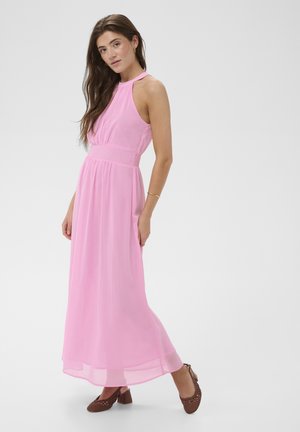 Nő ujjatlan rózsaszín maxi ruhában és barna szőtt magassarkú cipőben, egyik kezével oldalán áll egy egyszínű háttér előtt.