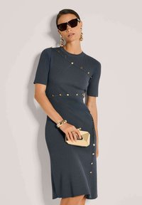 Frau in einem engen, dunkelgrauen Kleid mit goldenen Knöpfen, großen goldenen Ohrringen, schwarzen Sonnenbrillen, einem goldenen Armband und einer goldenen Clutch.