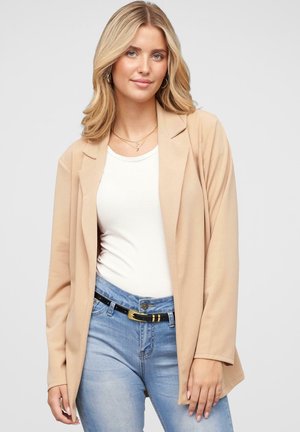Beige blazer med en glat tekstur, lange ærmer og en indsnittet krave, parret med en hvid top og lyseblå jeans, der har et sort bælte.
