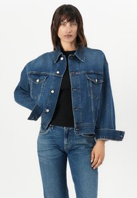 Veste en jean bleu avec une coupe ample, deux poches poitrine et des boutons en argent. Le tissu a une texture classique et un léger effet délavé.