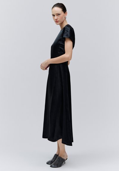 Femme aux cheveux foncés portant une robe midi noire élégante et des mules à talons cloutés noires, debout de profil devant un fond gris clair uni.