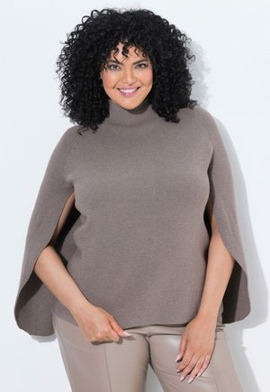 Kvinde med krøllet sort hår iført en taupe strikket kappe-trøje og beige læderbukser, smilende med hænderne hvilende nær hendes talje.