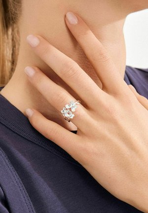 Hõbedast sõrmus lillemustriga, millel on mitu läbipaistvat vääriskivi, paigutatud õhukesele pandlale, kantud nimetissõrmel tumeda üleriide vastu.