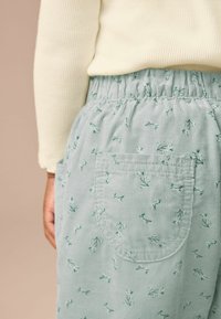 Hellblaue Cordhosen mit smocktem Bund und einer aufgesetzten Tasche, verziert mit kleinen grünen Blumenmustern.
