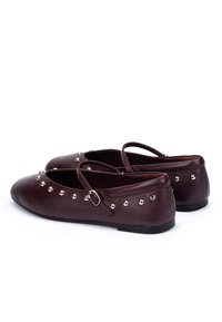 Mary Jane flat in pelle rossa scura con borchie argentate, cinturini con fibbia e suole nere piatte su sfondo bianco.