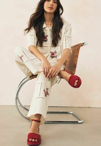 Witte geborduurde twee-delige outfit met bloemenpatronen, gecombineerd met rode suède plateau sandalen. Zittend op een gebogen metalen stoel.