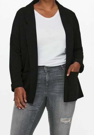 Personne portant une chemise blanche sous un blazer noir ouvert associée à un jean gris déchiré, main dans la poche du blazer, sur un fond uni.