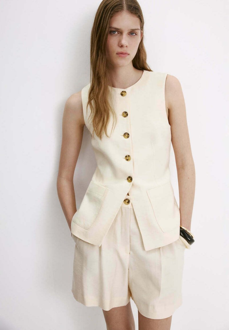 Jeune femme aux longs cheveux portant un gilet crème boutonné sans manches et un short taille haute assorti, les mains dans les poches, sur fond blanc.