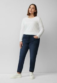 Weißes Langarmshirt, dunkelblaue Jeans und weiße Sneakers. Das Model steht selbstbewusst mit einer Hand in der Tasche vor einem schlichten Hintergrund.