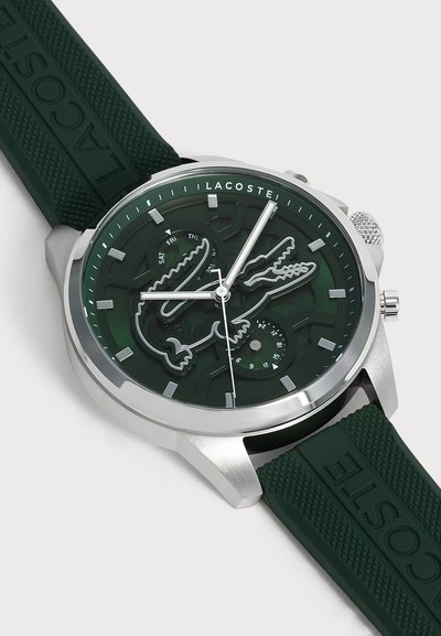 Часовник със зелен циферблат и каишка, сребърен корпус, лого на Lacoste и дизайн с крокодил, подциферблати за ден и дата, сребристи часови маркировки и стрелки.