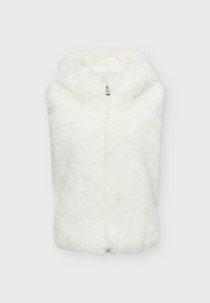 Un mannequin se tient devant un fond uni, vêtu d’un gilet en fourrure blanche par-dessus des vêtements noirs et de chaussures de ski noires.