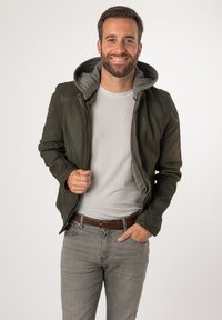 Olivgrüne Lederjacke mit grauem Kapuzen, taillierte Passform, halb offenem Reißverschluss, kombiniert mit einem hellgrauen T-Shirt und engen grauen Jeans.