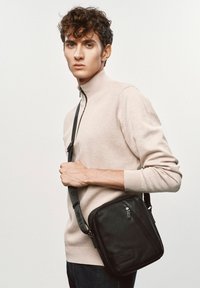 Czarna skórzana torebka na ramię o textured zakończeniu, z zapinaną na zamek przednią kieszenią i regulowanym paskiem. Nosi ją model w beżowym swetrze.