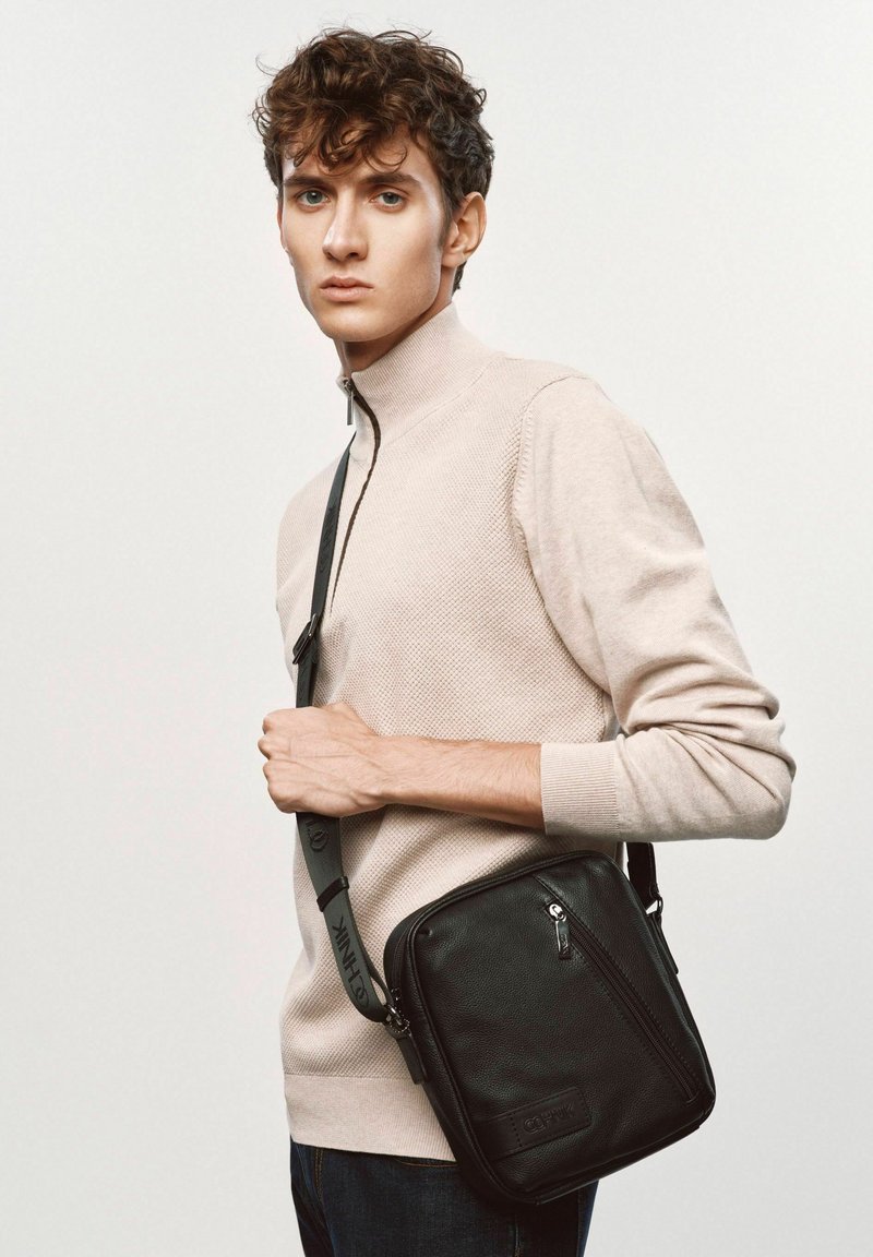 Czarna skórzana torebka na ramię o textured zakończeniu, z zapinaną na zamek przednią kieszenią i regulowanym paskiem. Nosi ją model w beżowym swetrze.