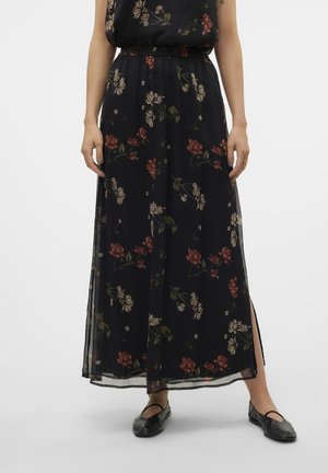 Kvinde iført en sort blomsterprint maxi nederdel med sidelommer og sorte balletsko, stående mod en ensfarvet hvid baggrund.