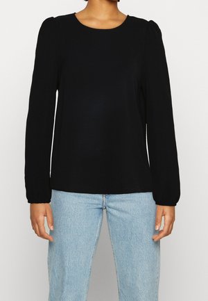 Nainen pukeutunut mustaan pitkähihainen toppiin, jossa on muhkeat hartiat, yhdistettynä vaaleansinisiin korkeavyötäröisiin farkkuihin, seisoo yksivärisen valkoisen taustan edessä.