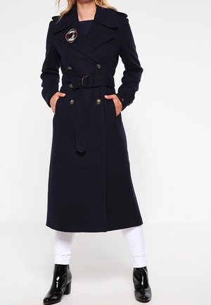 Kvinde iført en marineblå dobbeltradet frakke med bælte og en dekorativ broche, hvide bukser og sorte laklæder ankelstøvler.