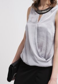 Haut sans manches argenté avec un col en V et un devant torsadé, orné d'un col en perles noires. Associé à une pochette noire.