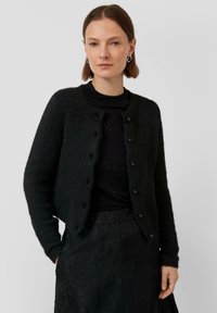 Frau mit kurzem braunen Haar, die silberne Creolen, eine schwarze, pl�üschige Knopfkardigan, ein schwarzes Top und einen strukturierten schwarzen Rock trägt.