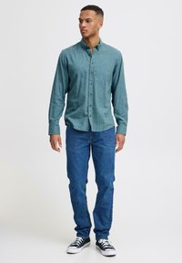 Chemise verte à boutons avec col, manches longues et ourlet arrondi ; associée à un jean bleu et des baskets noires.