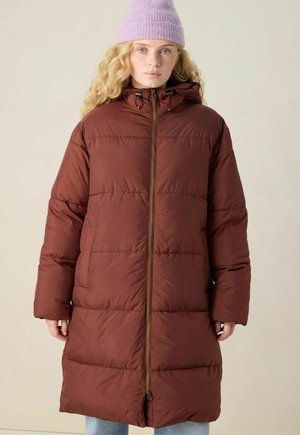 Young woman wearing a long brown puffer coat with hood and a light purple knit beanie, standing against a plain background.